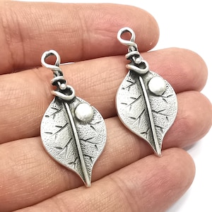 May include: Two silver leaf charms with a textured surface and a small round bead on each leaf. The charms have a wire loop at the top for attaching to jewelry.