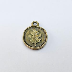 May include: A round, antique brass charm with a raised oak leaf design in the center. The charm has a small loop at the top for attaching to a necklace or bracelet. The outer edge is detailed with small, raised dots.