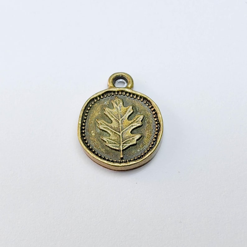 May include: A round, antique brass charm with a raised oak leaf design in the center. The charm has a small loop at the top for attaching to a necklace or bracelet. The outer edge is detailed with small, raised dots.