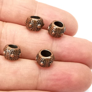 Peut inclure: Gros plan sur cinq petites perles métalliques de couleur cuivre. Chaque perle a une forme cylindrique avec des motifs décoratifs de points et de formes ovales. Les perles sont présentées sur fond blanc.