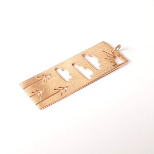 Puede incluir: Un amuleto de metal dorado con un diseño recortado de nubes y flechas. El amuleto tiene un bucle en la parte superior para sujetarlo a un collar o pulsera.
