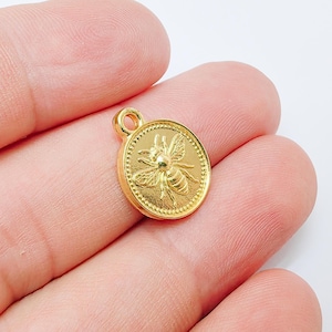 May include: A gold-colored round charm with a raised bee design. The charm has a small loop at the top for attaching to a necklace or bracelet. The bee is centered on the charm, surrounded by a dotted border.
