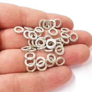 Puede incluir: Un montón de anillos de salto de plata, un tipo de hallazgo de joyería, en una mano. Los anillos son lisos y tienen una superficie ligeramente texturizada.