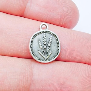 Könnte beinhalten: Ein kleiner, runder, silberfarbener Anhänger mit einem detaillierten Weizenähren-Design. Der Anhänger hat einen Perlenrand und eine Schlaufe oben zum Befestigen an Schmuck oder anderen Bastelarbeiten. Das Weizendesign ist auf dem Anhänger zentriert.