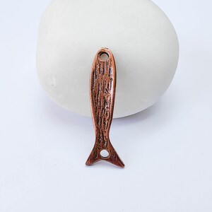 May include: A copper-colored fish-shaped pendant with a textured surface. The pendant has a hole at the top for hanging and a split tail. The fish pendant is displayed against a white background.