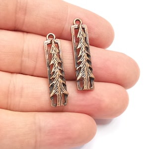 Könnte beinhalten: Zwei kupferfarbene, rechteckige Anhänger mit einem ausgeschnittenen Baumdesign. Jeder Anhänger hat oben eine kleine Schlaufe zum Befestigen an Schmuck. Das Baumdesign ist detailliert mit sichtbaren Ästen und Nadeln.