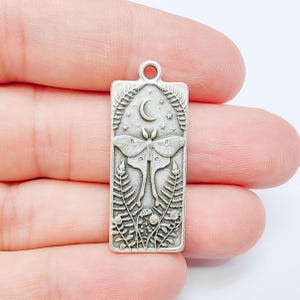 May include: A rectangular silver-toned metal pendant featuring an embossed design of a moth centered between two fern fronds. Above the moth is a crescent moon surrounded by small stars. The pendant has a small loop at the top for attachment.