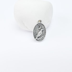 May include: Silver charm featuring a detailed design of a crow perched on a branch with a crescent moon above it.