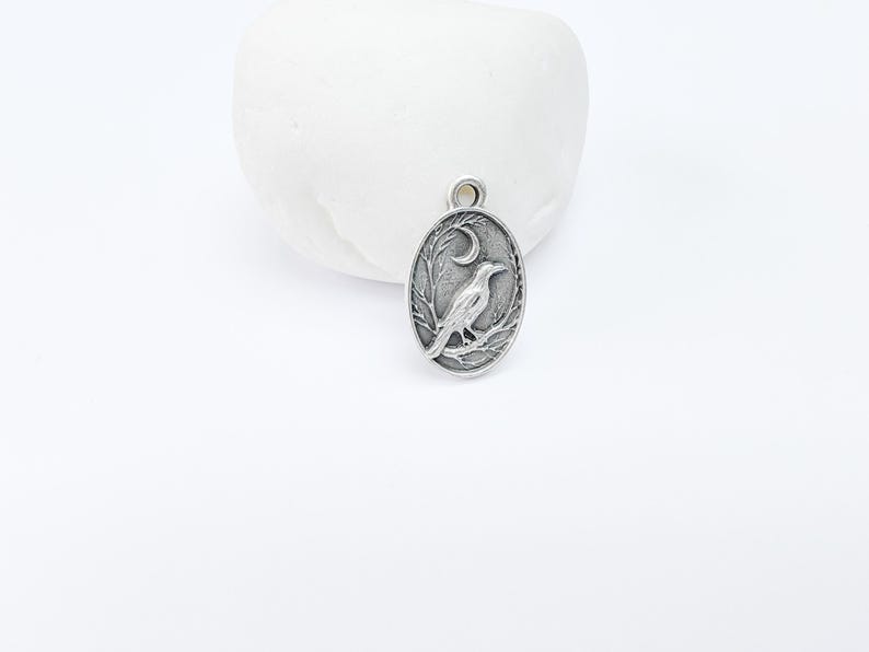 May include: Silver charm featuring a detailed design of a crow perched on a branch with a crescent moon above it.
