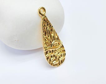 May include: A gold-colored pendant with a textured surface and a teardrop shape. The pendant features an engraved design of a sun, eye, and abstract shapes. It has a small loop at the top for attaching to a chain or cord.
