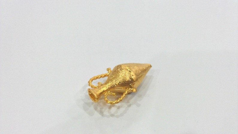 May include: A gold-colored metal pendant in the shape of a small, ornate vase with a pointed top.