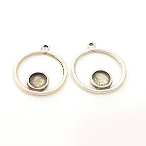 May include: Two silver-coloured pendant blanks. Each has a circular frame with a small, round bezel in the centre. These pendants are designed for jewellery making, with a loop at the top for attaching to a chain or cord. The background is plain white.