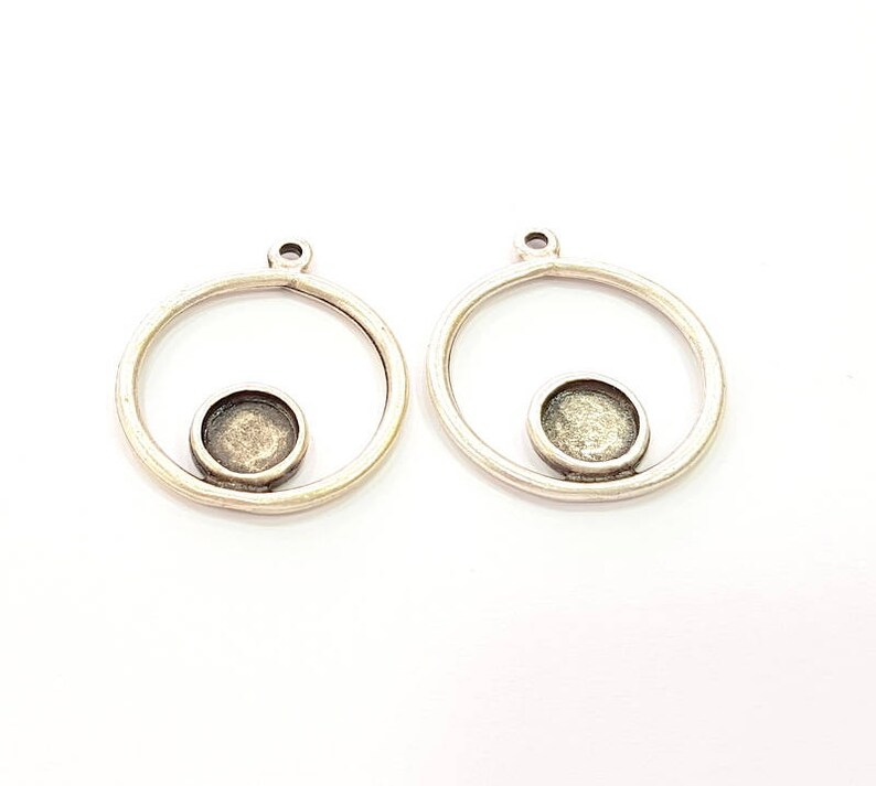 May include: Two silver-coloured pendant blanks. Each has a circular frame with a small, round bezel in the centre. These pendants are designed for jewellery making, with a loop at the top for attaching to a chain or cord. The background is plain white.