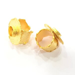 Op de afbeelding: Twee goudkleurige ringen met een getextureerd oppervlak en een uniek ontwerp. Eén ring heeft een cirkelvorm met een verhoogd midden, de andere heeft een vergelijkbaar ontwerp met een bloembladachtige accent. De ringen staan tegen een witte achtergrond.