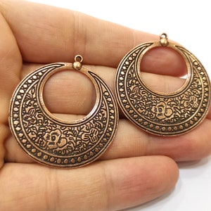May include: Two copper-colored, circular pendants with floral and dot patterns. Each pendant has a small loop at the top for hanging. The pendants are likely for jewelry making or crafting.