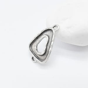 Puede incluir: Pendiente de metal plateado con forma triangular y un pequeño recorte en forma de lágrima en el centro. El pendiente tiene una superficie texturizada y un bucle en la parte superior para sujetarlo a una cadena o cordón.