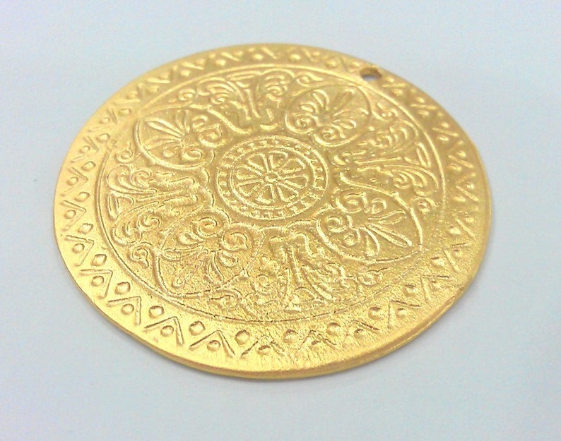Peut inclure: Un pendentif en m&eacute;tal dor&eacute; avec un motif en relief complexe. Le pendentif est rond et poss&egrave;de un trou en haut pour l'accrocher.