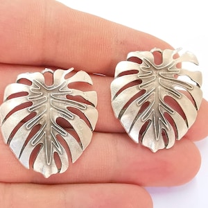 May include: Two silver-toned metal charms shaped like monstera leaves. The leaves have a detailed cut-out design.