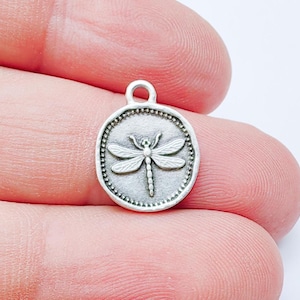 May include: A silver-toned dragonfly charm with a detailed insect design. The charm is round with a beaded border and a loop for attaching to jewelry. The dragonfly has detailed wings and body.
