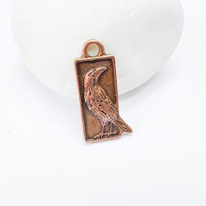 May include: A copper-coloured charm featuring a detailed, embossed raven in a rectangular frame. The charm has a loop at the top for attaching to jewellery or other crafts.