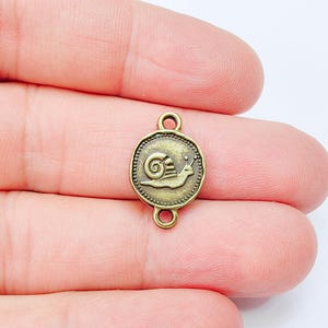 Pode incluir: Um berloque de metal bronzeado com um desenho de caracol. O berloque redondo tem uma borda com contas e duas argolas para fixação. O caracol é retratado em perfil, ideal para criar joias.