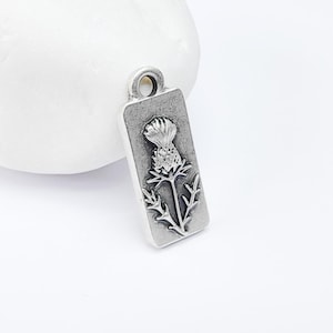 Może przedstawiać: Srebrny charm z detalicznym, tłoczonym obrazem kwiatu ostu.