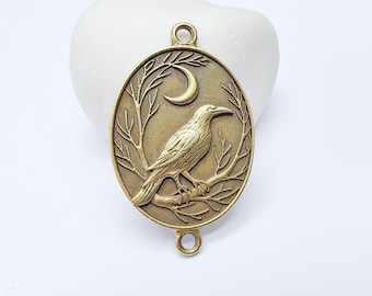Op de afbeelding: Antiek messingkleurige ovale hanger met een gedetailleerd reliëf van een raaf die op een tak zit, tegen een halve maan en boomtakken. De hanger heeft twee lussen om aan een ketting of andere sieradenonderdelen te bevestigen.
