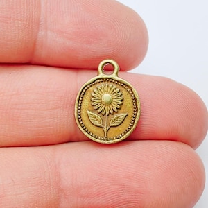 May include: A small, antique gold-coloured charm featuring a sunflower design. The charm is round with a beaded border and a loop at the top for attachment. The sunflower is centrally placed, with detailed petals and leaves, creating a vintage aesthetic.