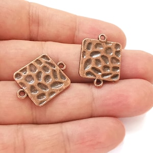 Op de afbeelding: Twee vierkante connectorbedels van koperkleurig metaal met een getextureerd oppervlak. Elke bedel heeft twee springringen.