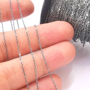 May include: A close-up of a silver chain, showing the chain's texture and detail. The chain is lying on a hand, with a spool of the same chain in the background.