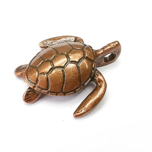 Puede incluir: Un pequeño amuleto de tortuga de color cobre con una concha detallada y un bucle para sujetarlo a un collar o pulsera.