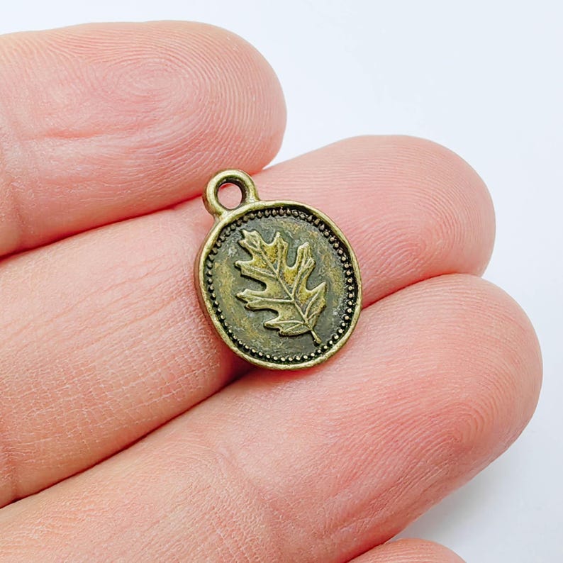 May include: A small, round, antique brass-colored charm with a raised oak leaf design in the center. The charm has a beaded border and a small loop at the top for attaching to a chain or bracelet. Ideal for jewelry making.