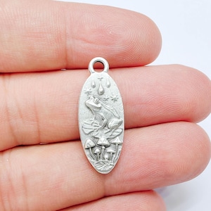 May include: An oval-shaped silver-toned metal charm featuring a raised relief design of a frog sitting on a lily pad, surrounded by several small mushrooms at the base and raindrops with stars depicted in the upper section, including a top loop.