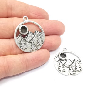 May include: Two silver-toned metal charms in the shape of a circle with a mountain scene and a sun cutout. The charms have a textured surface and are perfect for jewelry making.