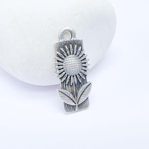 May include: Silver charm featuring a sunflower with leaves, designed to look like a wood carving. The charm has a loop at the top for attaching to a necklace or bracelet.