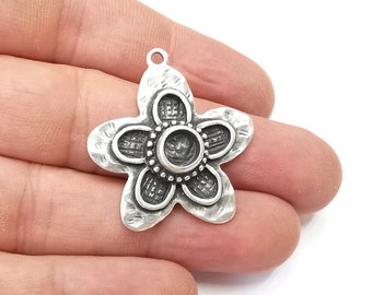 Puede incluir: Un colgante plateado con forma de flor y superficie texturizada. El colgante tiene cinco pétalos y un engaste circular central. Tiene un pequeño lazo en la parte superior para sujetarlo a una cadena u otro componente de joyería.
