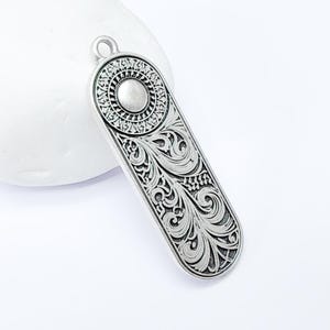 Op de afbeelding: Zilveren hanger met een sierlijk ontwerp met een cirkelvormig midden met een verhoogd, zilverkleurig punt. De hanger heeft een lus aan de bovenkant om een ​​ketting aan te bevestigen.