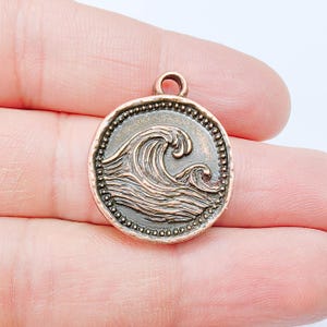 Pode incluir: Um berloque redondo, cor de bronze, com um design detalhado de ondas. O berloque apresenta um padrão de ondas em relevo dentro de uma borda de contas e uma pequena argola na parte superior para fixação. Estética vintage e náutica.