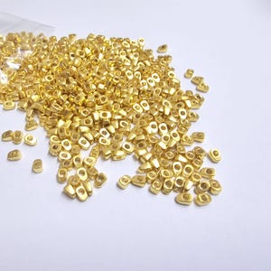 May include: A collection of small, gold-colored metal beads scattered on a white surface. The beads are oval-shaped with a hole in the center, and some are still in a clear plastic bag. The beads have a shiny finish.