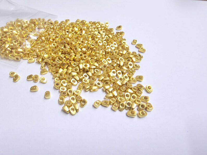 May include: A collection of small, gold-colored metal beads scattered on a white surface. The beads are oval-shaped with a hole in the center, and some are still in a clear plastic bag. The beads have a shiny finish.