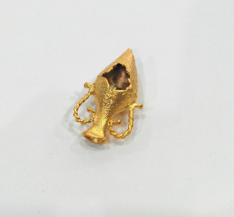 May include: A gold-colored metal pendant shaped like a small, ornate vase with a broken top. The vase has a textured surface and intricate details.