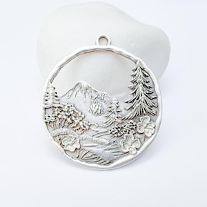 May include: A silver-colored pendant with a mountain and forest scene. The round pendant features a detailed relief of a mountain, trees, and flowers within a circular frame. It has a small loop at the top for hanging.