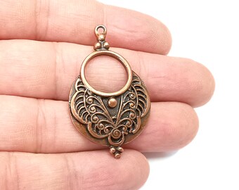 Op de afbeelding: Een koperkleurige hanger met een ingewikkeld, filigraan ontwerp. De hanger heeft een ronde opening in het midden en een afgeronde onderkant.