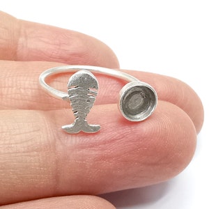 May include: A silver-colored ring with a fish-shaped design and a small, round setting. The fish has textured details, and the ring band curves around to the setting. The ring is held in a hand against a white background.