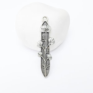 Op de afbeelding: Een zilverkleurige hanger met een bloemmotief. De hanger heeft een centrale stengel met gedetailleerde bladeren en bloemachtige vormen. Hij heeft een lus aan de bovenkant om aan een ketting of koord te bevestigen. De hanger is waarschijnlijk van metaal.