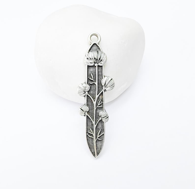 Op de afbeelding: Een zilverkleurige hanger met een bloemmotief. De hanger heeft een centrale stengel met gedetailleerde bladeren en bloemachtige vormen. Hij heeft een lus aan de bovenkant om aan een ketting of koord te bevestigen. De hanger is waarschijnlijk van metaal.