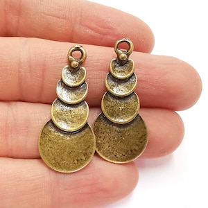 Peut inclure: Une paire de breloques de boucles d'oreilles en métal doré antique avec un design superposé en cascade. Chaque breloque présente cinq disques ronds empilés les uns sur les autres, créant un effet étagé.