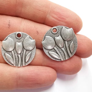 May include: Two silver-toned metal charms in the shape of a circle with three tulips embossed on each charm. Each charm has a hole at the top for attaching to a necklace or bracelet.