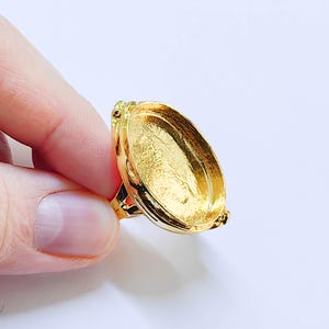 Op de afbeelding: Een goudkleurige ring met een ovale zetting. De ring heeft een gedetailleerd ontwerp rond de zetting en band. De zetting is leeg, klaar voor een edelsteen of andere versiering. De ring wordt door een persoon vastgehouden.