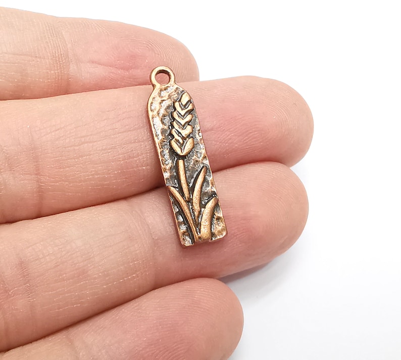 May include: A copper-toned metal charm with a wheat stalk design. The charm has a textured surface and a loop at the top for attaching to a necklace or bracelet.